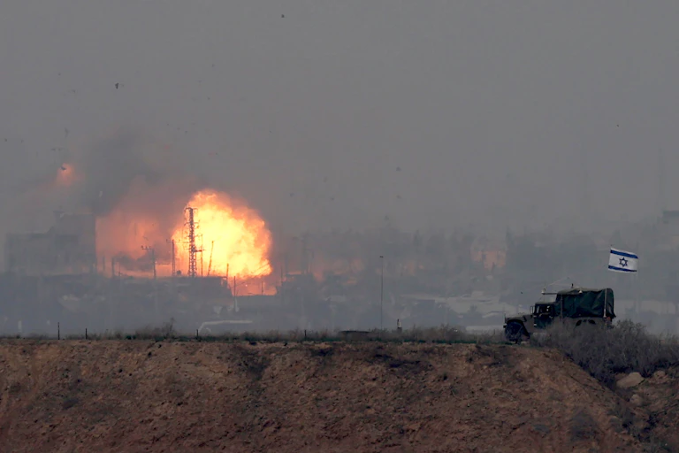 An explosion in a city in the background with a military vehicle flying an Israeli flag in the foreground.