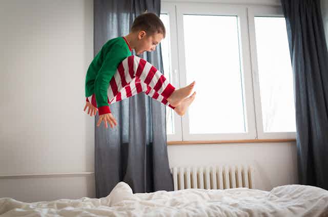 A boy jumps on the bed while wearing Christmas-themed pyjamas.