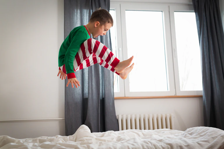 A boy jumps on the bed while wearing Christmas-themed pyjamas.