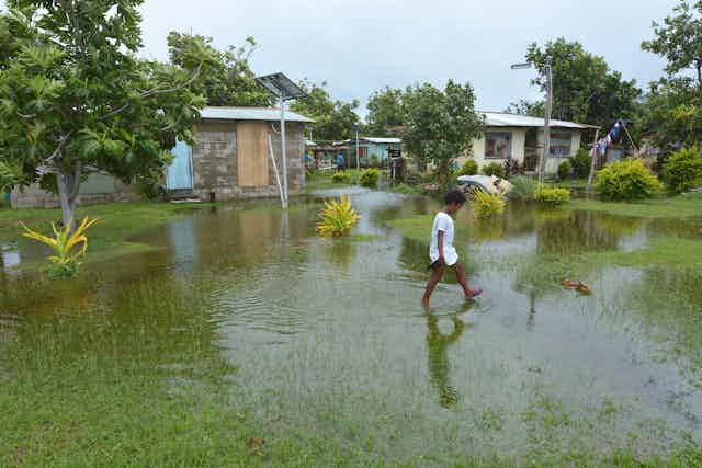 A small girl walking through water on a green patch in front of some houses