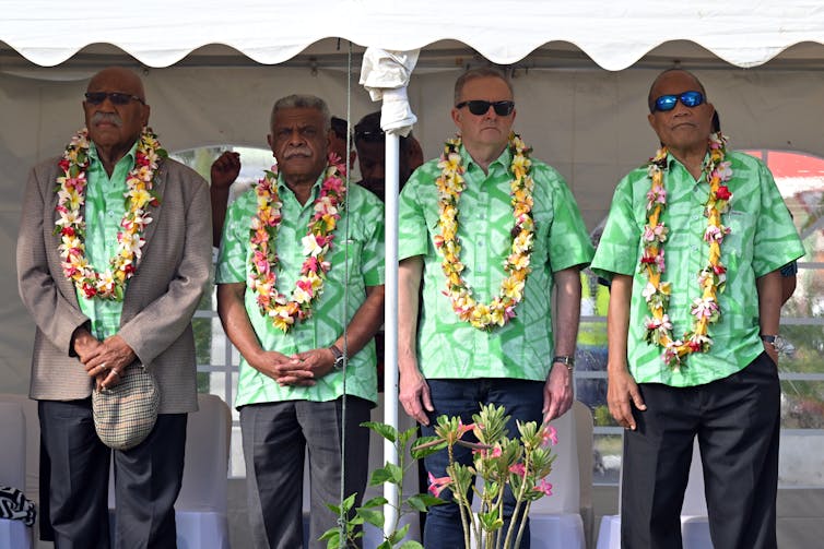 Four men in green short sleeve shirts with leis around their necks standing on a verandah