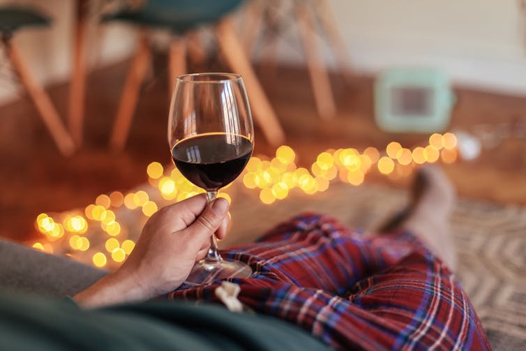 Man sitting on sofa, wearing pyjamas, holding glass of red wine, sparkling lights on floor