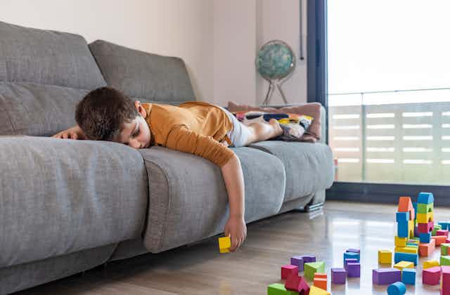 A bored child lies down on a couch while listlessly playing with blocks.