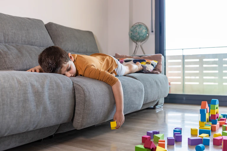 A bored child lies down on a couch while listlessly playing with blocks.