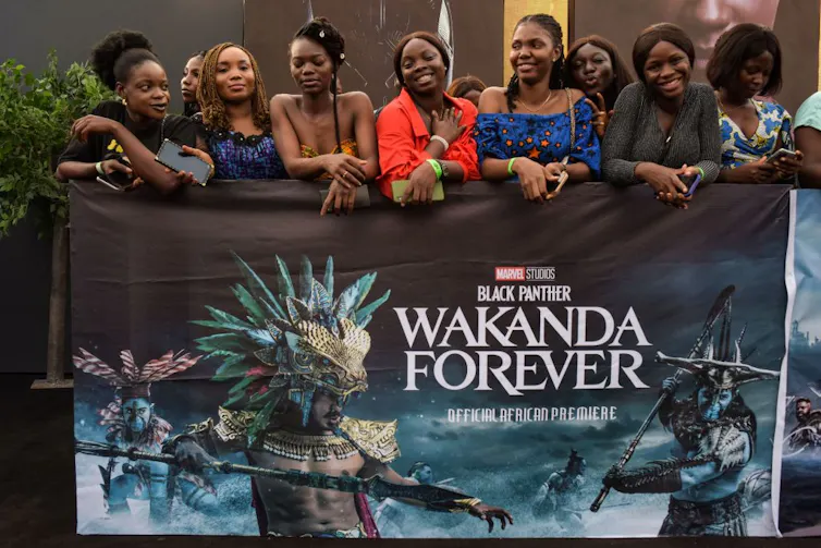 Seven smiling and smartly dressed women lean against a metal bar with a poster hanging from it reading