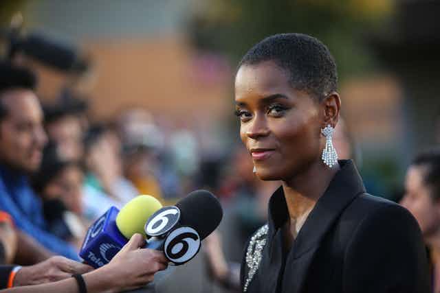 A woman with short natural hair and a black jacket with silver adornment glances at the camera with a small smile. In front of her a row of people, some holding microphones to interview her.