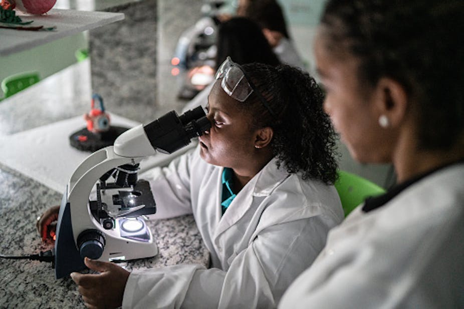A woman in a white coat looks through a microscope while another woman dressed similarly looks at her