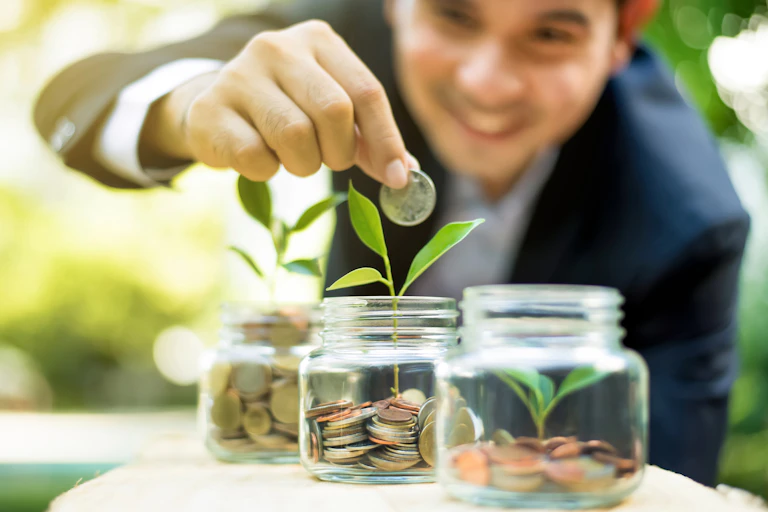 Man in suit putting coins in jars with green shoots growing out of them.