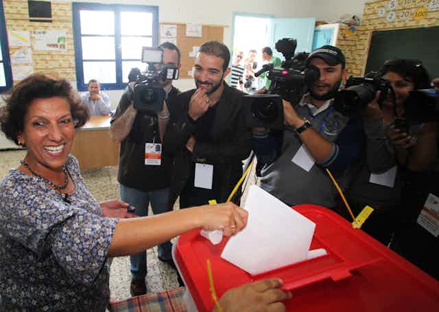 A smiling woman casts a vote while press cameras film her. 