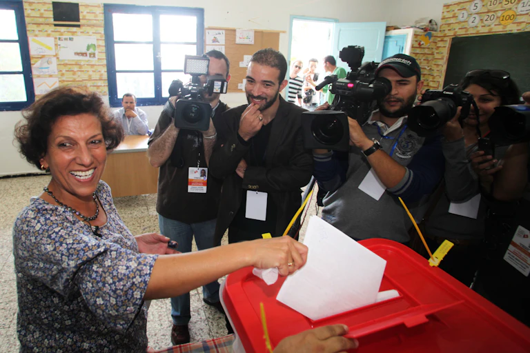 A smiling woman casts a vote while press cameras film her.