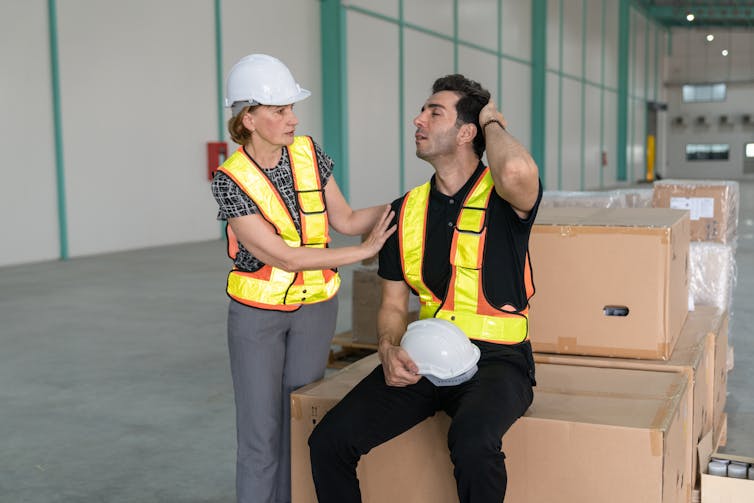 Manager in high-vis vest and hard hat comforts team member in high-vis vest, sitting on boxes in a warehouse.