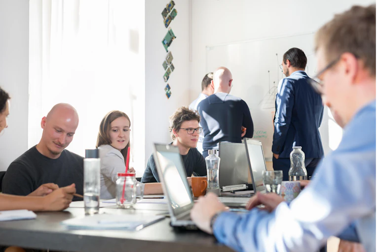 A group of people sit around a large desk with laptops having an informal meeting