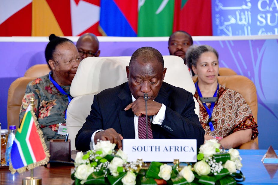 A man in a suit and tie sits in front of a microphone, with a label on the table saying 'South Africa'.
