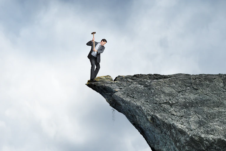 A businessman swings a sledge hammer against a rocky cliff as he stands on the wrong side placing himself at great risk.