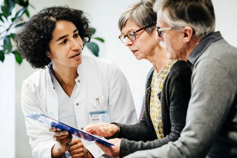 A doctor in a white coat, with thick black hair, looks thoughtfully at two seated patients with glasses as they study something on a clipboard.