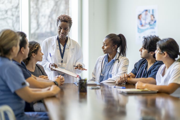 Six young adults sit at a conference table, some of them in scrubs, as a doctor in a white coat leads a discussion.