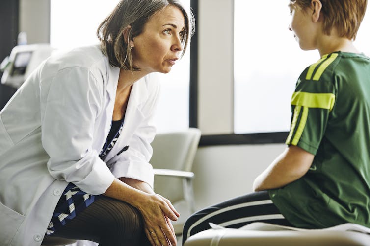 A woman in a white medical coat leans forward, seated, as she talks seriously with a seated boy in a green t-shirt.