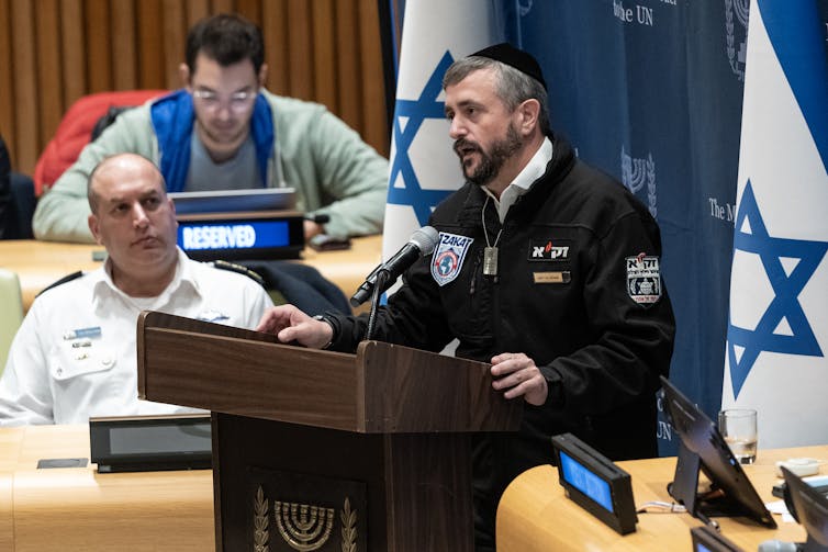 A man wearing a dark jacket with Hebrew words on it and a kippah stands at a wooden podium and speaks to people seated. He stands in between two Israel flags.