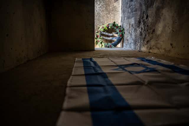 An Israeli flag is draped on the floor of a stone outdoor enclosure, with a wreath with Hebrew letters seen at the edge of one wall. 
