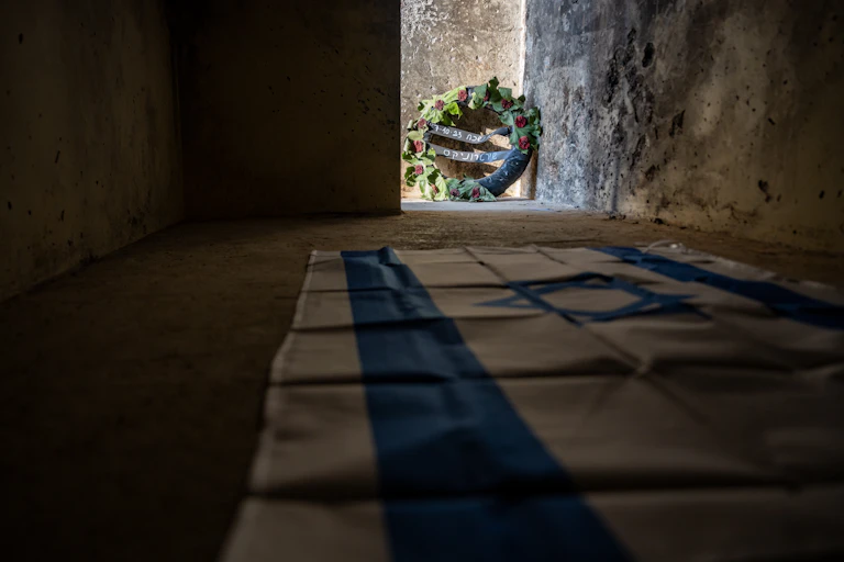 An Israeli flag is draped on the floor of a stone outdoor enclosure, with a wreath with Hebrew letters seen at the edge of one wall.