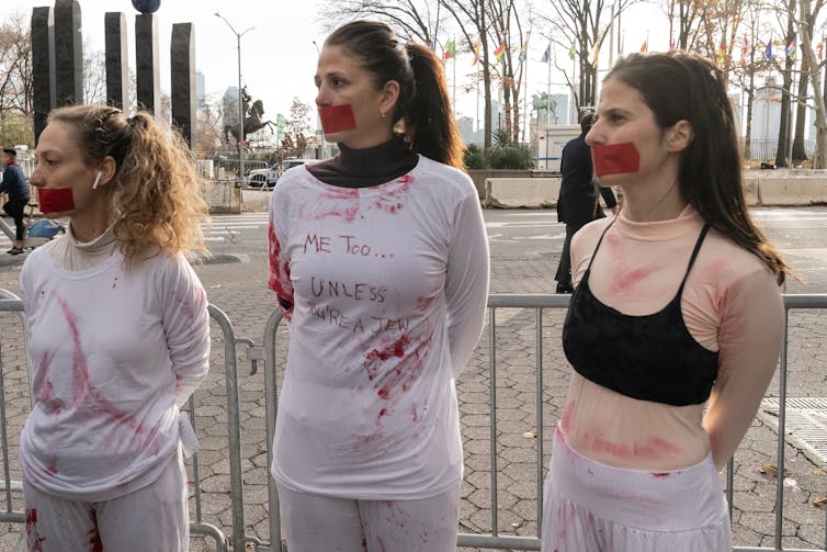 Three young women stand with red tape covering their mouths. Two wear white clothing that appears stained with red coloring and one has the words 'me too.. unless you're a Jew.' They stand across the street from the UN in New York City.