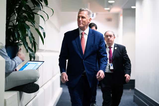 Kevin McCarthy wears a dark blue suit and a pink tie as he walks down a hallway, with two men in suits behind him. 