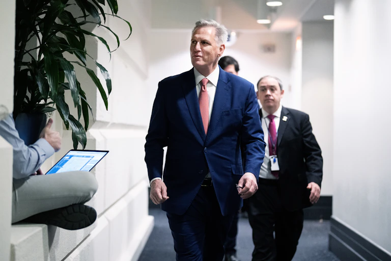 Kevin McCarthy wears a dark blue suit and a pink tie as he walks down a hallway, with two men in suits behind him.