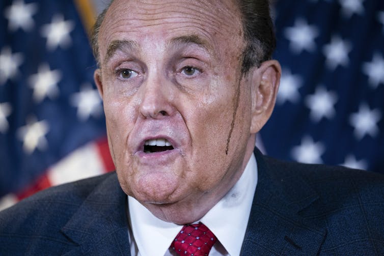 Rudy Giuliani appears to speak with his mouth dropped open, and American flags behind him. He has dark dye running down his face.