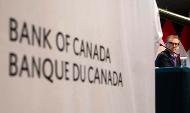 A man behind a microphone sits next to a sign that reads Bank of Canada/Banque du Canada.