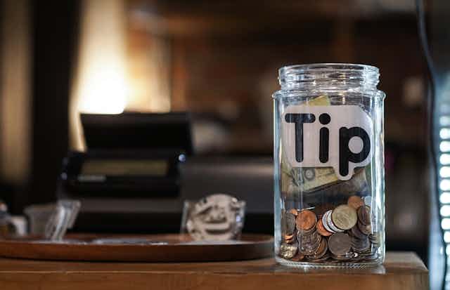 A glass jar full of money with a 'tip' label sits on a checkout counter