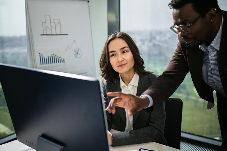 man pointing at computer screen with seated woman looking on