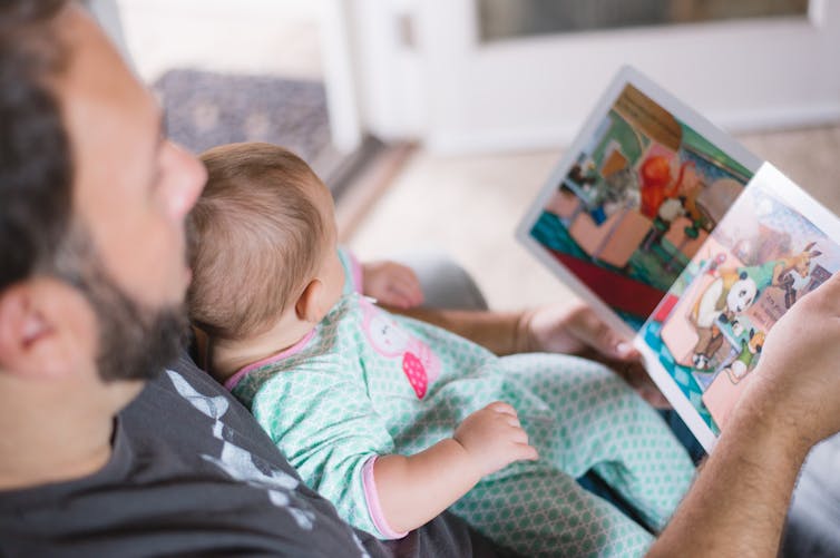 A baby in a mint-green sleeper sits in her father's lap while he reads her a book.