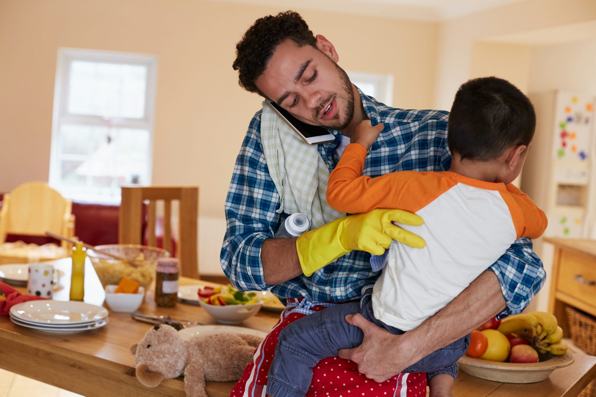 A man talks into his cellphone while holding a child. He has a cleaning glove on one hand and there's a baby bottle under his arm.