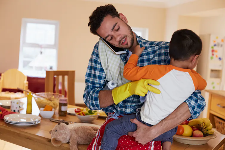 A man talks into his cellphone while holding a child. He has a cleaning glove on one hand and there's a baby bottle under his arm.