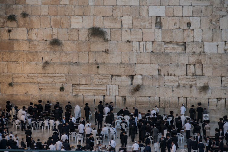 A large group of men in black and white clothing, seen from the back, face a tall tan-colored stone wall.