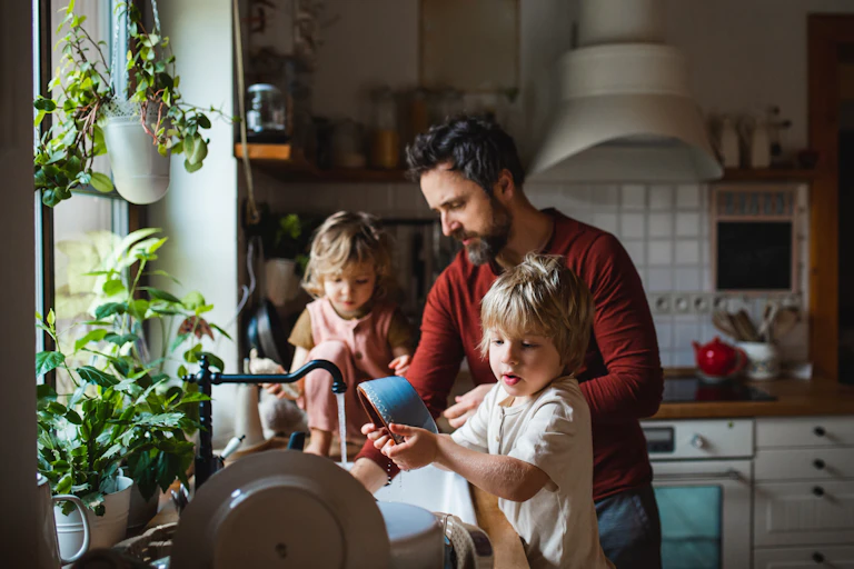 A bearded man washes dishes with two small children.
