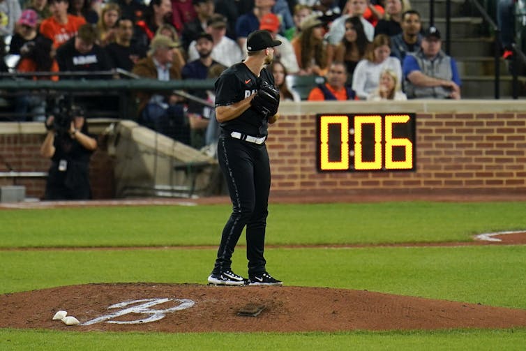 A pitcher in a black baseball uniform stands on a pitcher's mound. In the background, a digital timer displays 0:06.