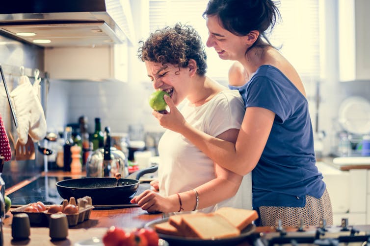 Same-sex female couple cooking in kitchen, one feeding the other fruit