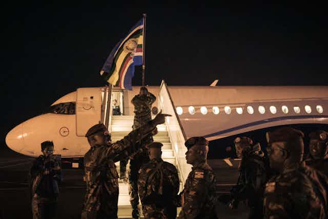 A man in military fatigues holding up a flag as he climbs up the steps towards a plane with several other men dressed similarly waiting to board