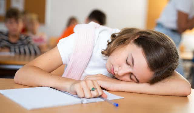 An adolescent girl sleeps on her desk with a pen in her hand and her notebook open.