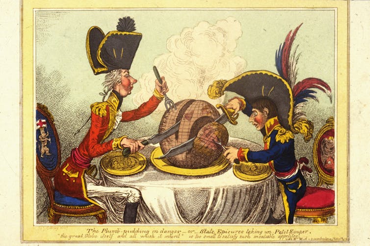 Two men in military uniforms with large hats cutting a large, round, brown pudding, placed on a table between them.