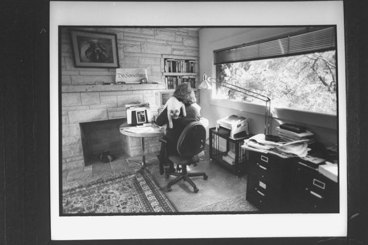 A woman working in a home office at her desk with a cat on her shoulder, seen from her back.