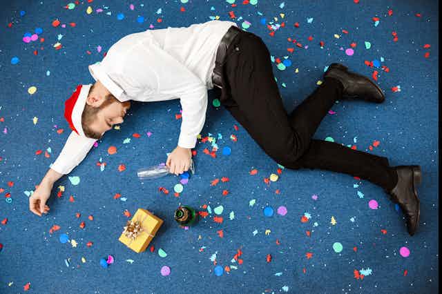 Man in shirt and trousers and santa hat with empty glass, lying on the floor surrounded by confetti, a present and a champagne bottle.