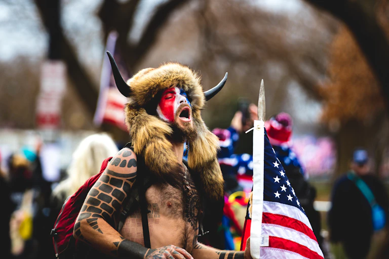 A topless Jacob Chansley with a painted face and horned headress holding an American flag.