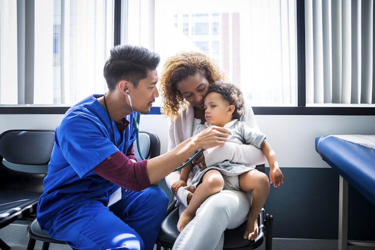 Clinician holding stethoscope over the chest of a toddler sitting in the lap of their parent