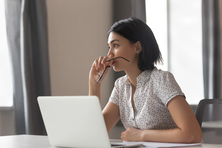 An Asian woman stares off to the side while sitting in front of an open laptop