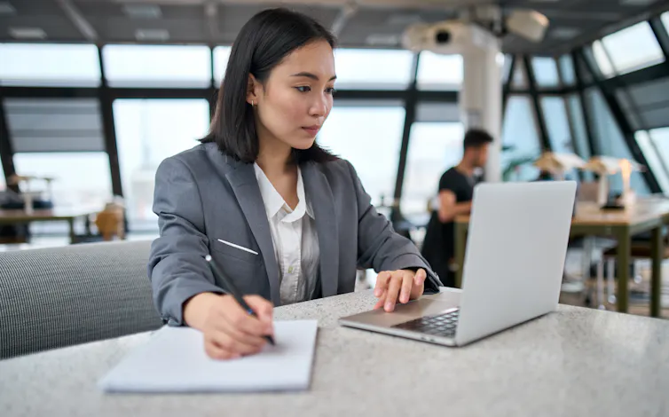 An Asian woman in business attire working on a laptop at a desk in a communal office space