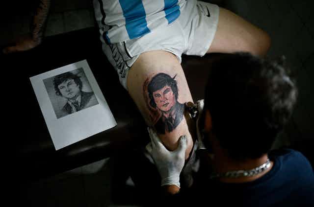 Tatoueur en train de tatouer le visage de Javier Milei sur la cuisse d'un. homme en maillot de l'équipe nationale de football d'Argentine