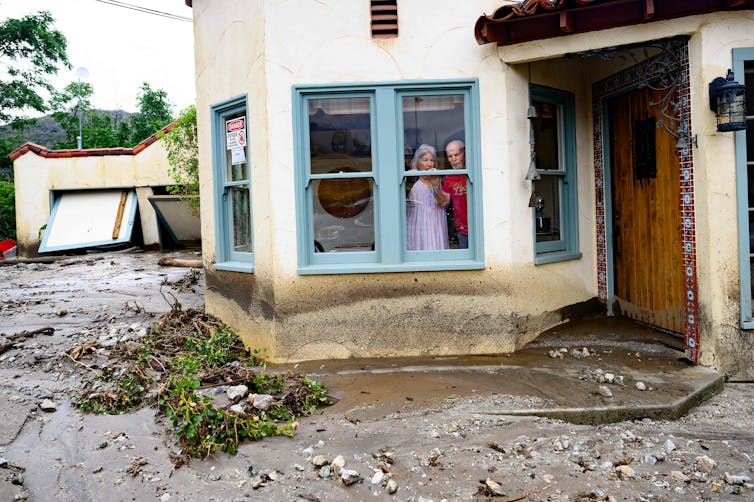 Two older adults look out a window over a yard turned to mud. The mudline on the house is almost up to the window sill, and the garage's doors have been torn off and are leaning down.