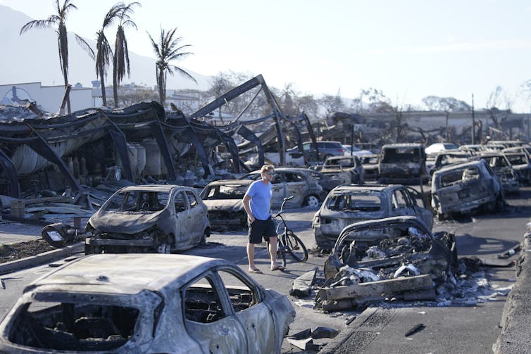 A man in shorts in flipflops walks among burned out cars. Not much remains of the houses.